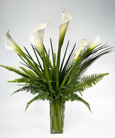Bouquet of white calla lilies in a clear glass vase with greenery.